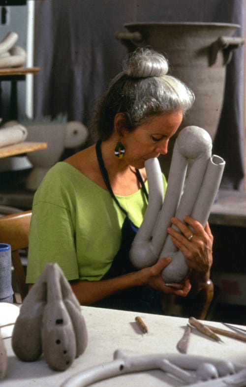 Image:&nbsp;Ms. Rawcliffe in her studio; in the foreground, there is a triple ocarina and a round ceremonial multiperson instrument with whistles &amp; flutes. In the background are the Whistling Pot for 3, and a two person chamber duct piece, After…