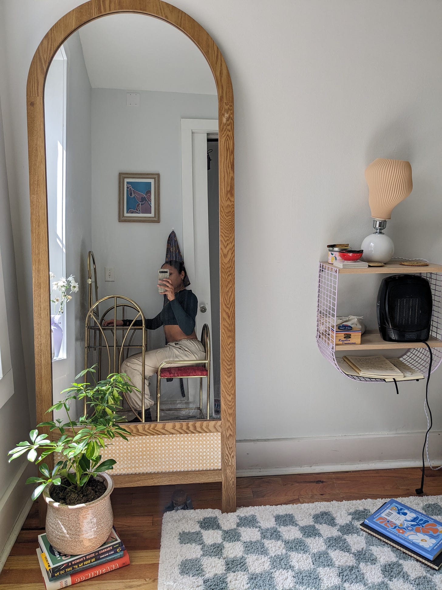 Nathalie sits by her gold vanity. She takes a photo of herself towards her full length mirror across her bedroom. Her seafoam green and white checkered rug peeks through