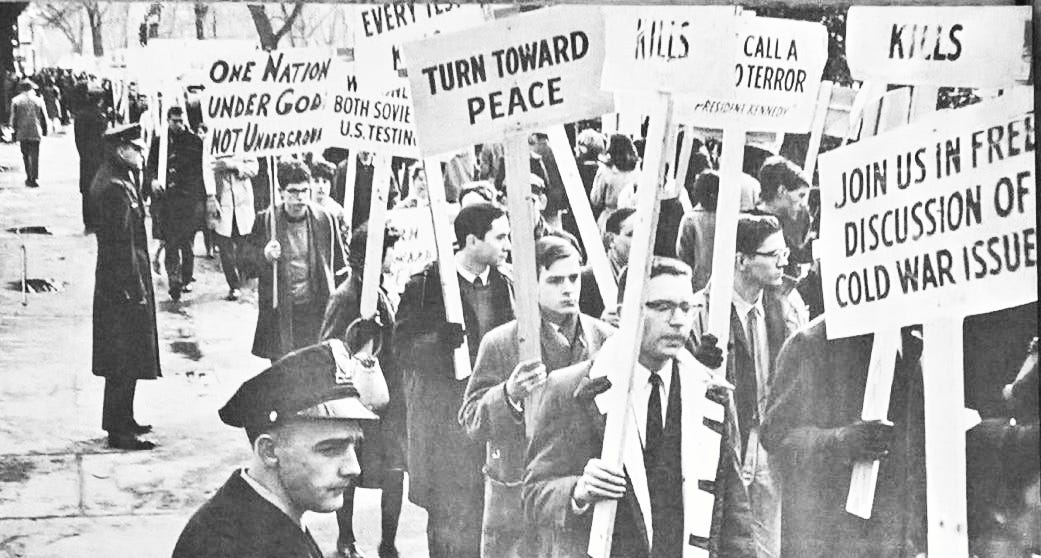 Black and white photograph of young people protesting in Washington, DC, while polite look on with polite disinterest. The signs carried by the protesters advocate for peace, against nuclear testing, and against fallout shelters.