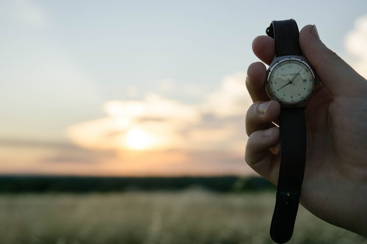 Photo of a person holding an analog watch in front of a field with the sun coming up in the distance. Photo of a person holding an analog watch in front of a field with the sun coming up in the distance.