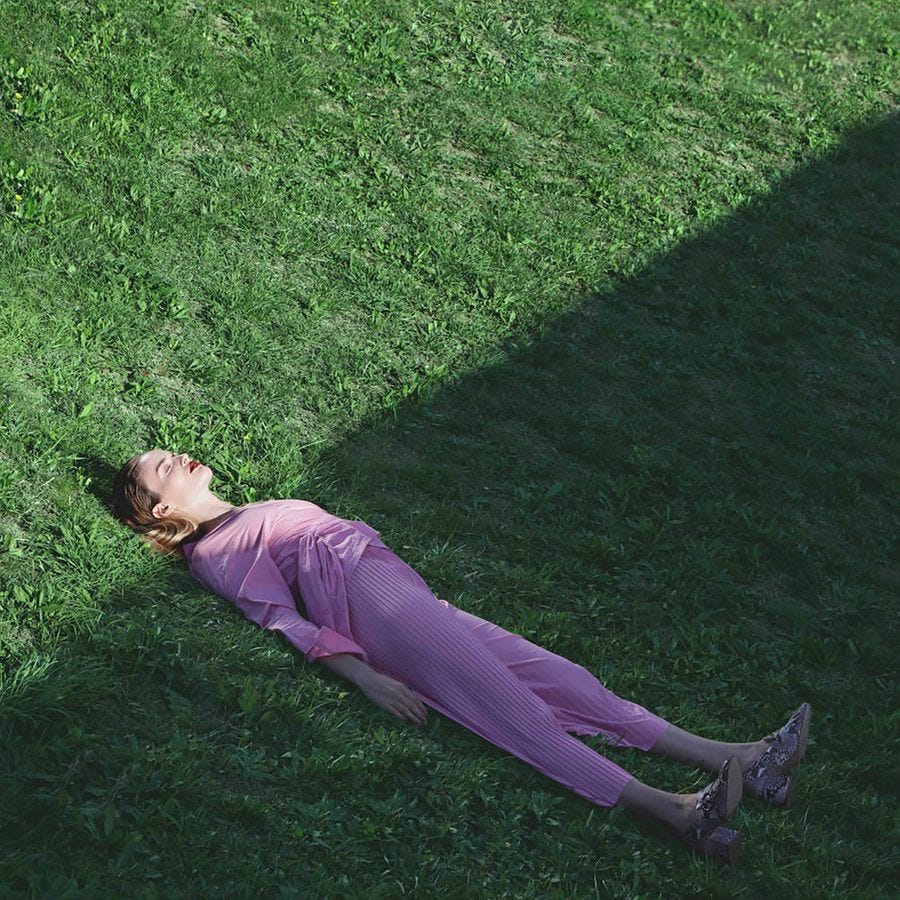 A woman wearing a lilac coloured trouser suit and with eyes closed, lies flat on their back on a a green grassy space.
