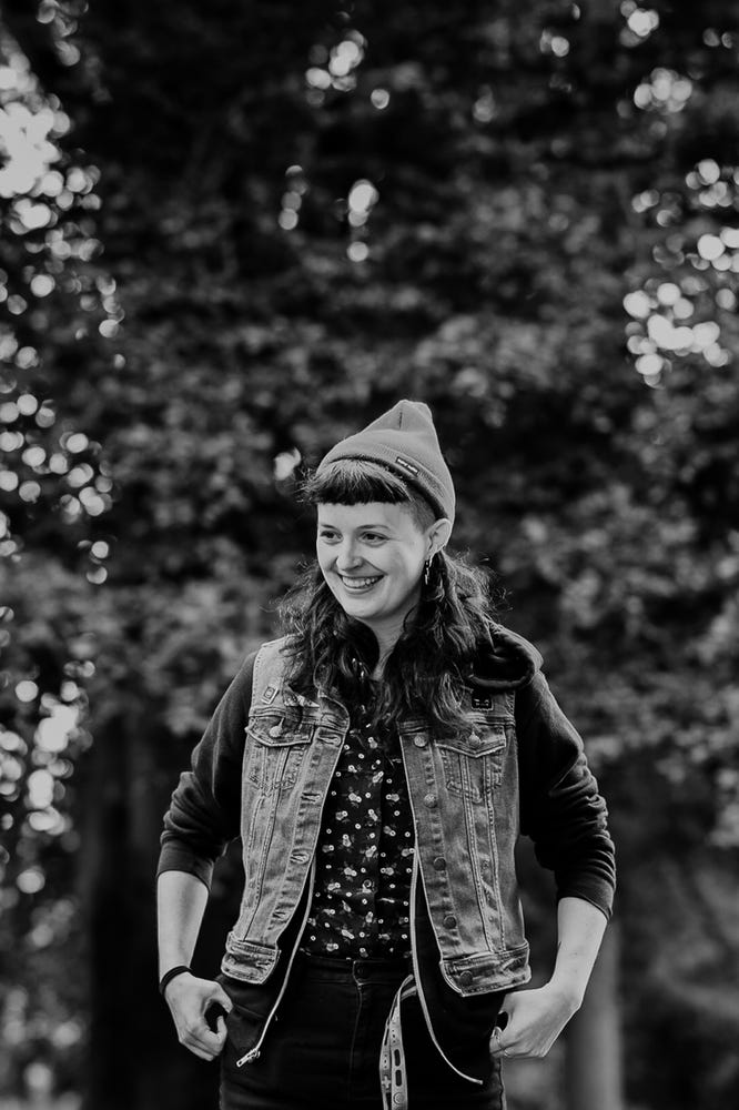 B+W photo of a person identifying as Queer (they/them) in a red beanie hat, brown cropped bangs, hands holding front of jean vest with black shirt and black pants, slight smile on their face, standing in front of a brick wall