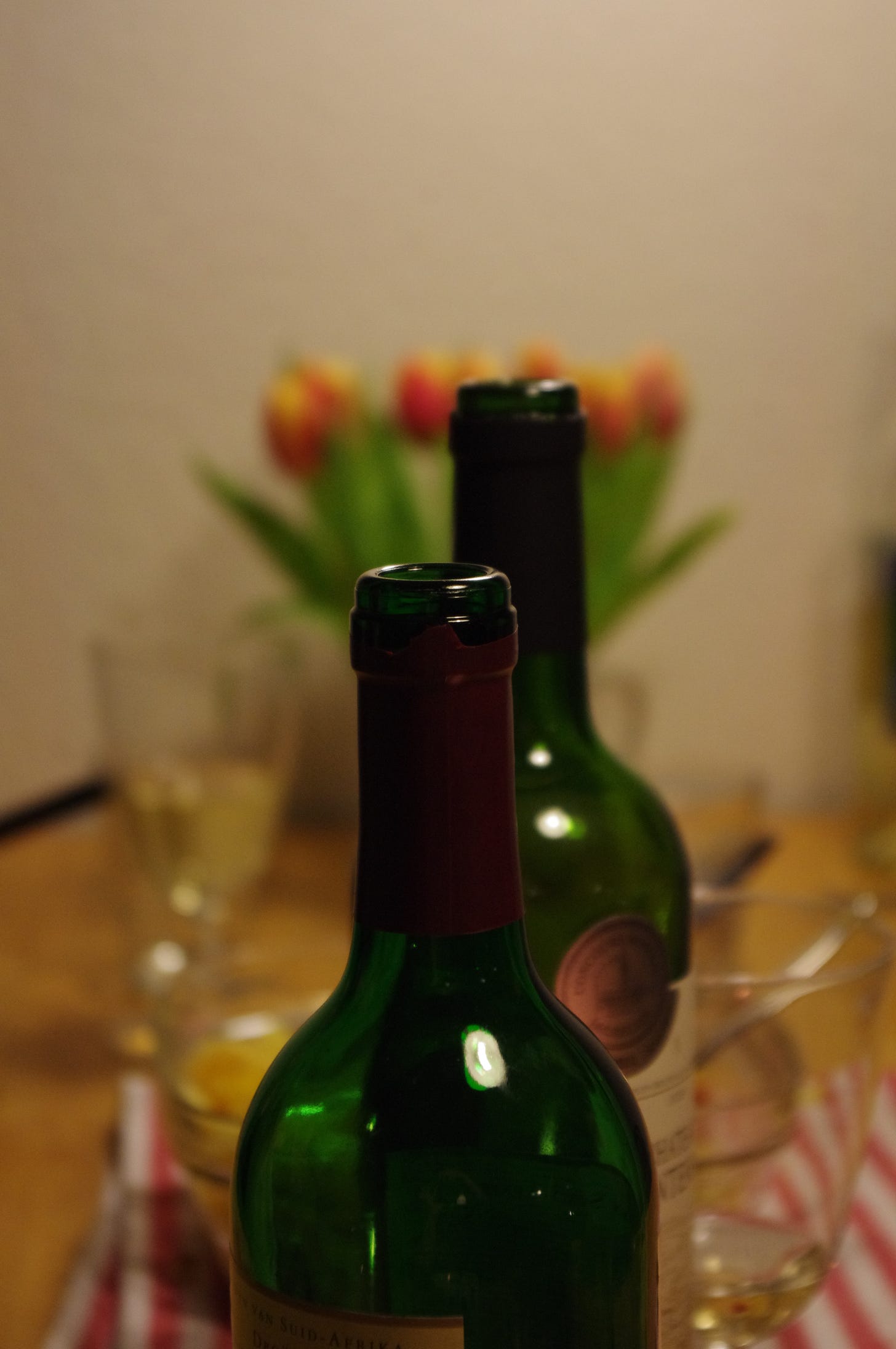zwei offene Weinflaschen, im Hintergrund ein verschwommener Strauß Tulpen und mehrere Gläser