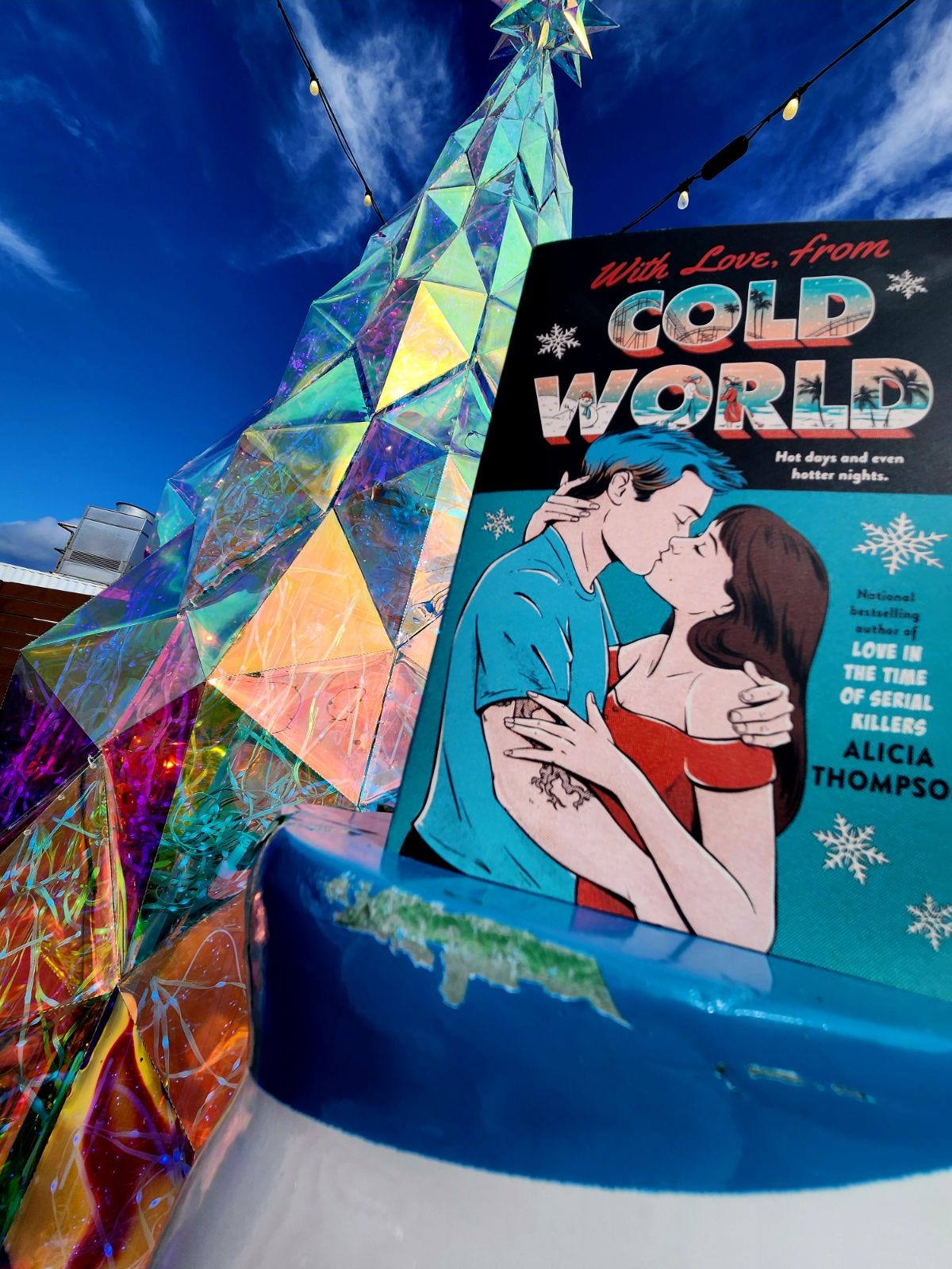 A copy of the teal cover copy of WITH LOVE, FROM COLD WORLD in front of an iridescent Christmas tree