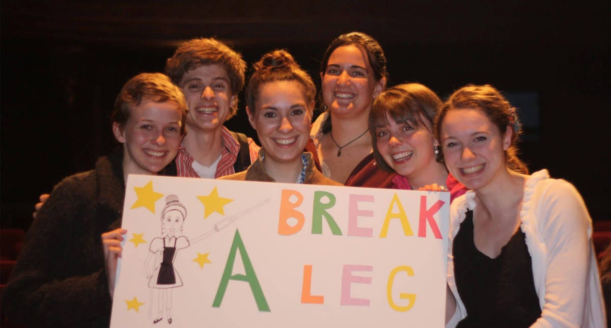 Shell stands in the middle of five other friends. They are all holding a handmade sign that says Break a Leg with a drawing of Shell in character as Berta on it.