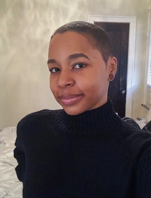 Black woman with buzzed short black hair in a selfie with a smile on in a bed room in a black turtle neck.