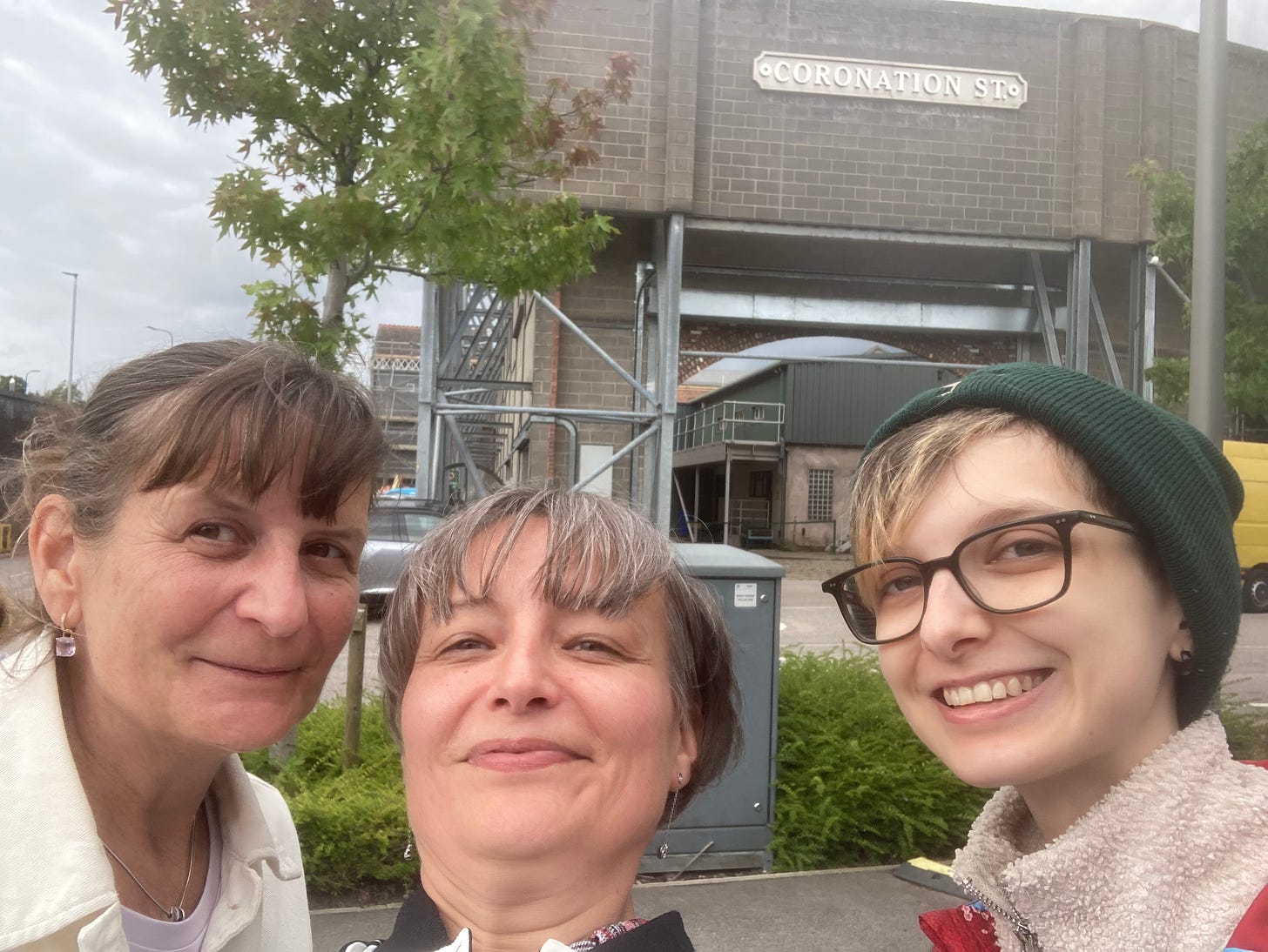 Selfie of Beck's mum, Beck's auntie and Beck in front of scaffolding with a big Coronation Street sign on a brick facade. Beck's mum is a white woman with a brown bob, tied up in a pony tail. Beck's auntie is a white woman with cropped grey hair, and Beck is an androgynous white person wearing square glasses and a green beanie with their blond and brown highlighted fringe peaking out.