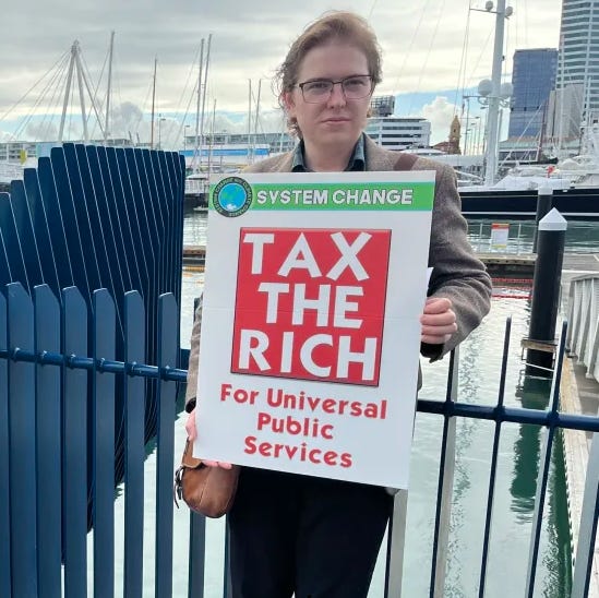 Image depicts the author at a protest in Auckland holding a sign which reads "Tax the Rich for Universal Public Services."