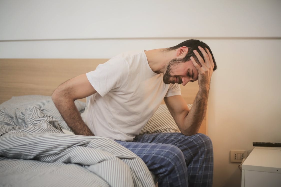 Free A young man in pajamas holding his head, sitting on a bed, appears to be experiencing a headache. Stock Photo