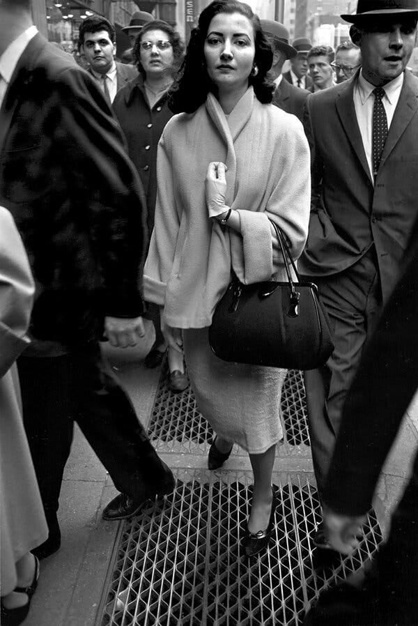 A black-and-white photo of a street scene with a crowd of people walking on a sidewalk, mostly in dark suits, and in the center is a woman in a light-colored suit, staring straight ahead, one arm curled up to her chest holding a black purse.