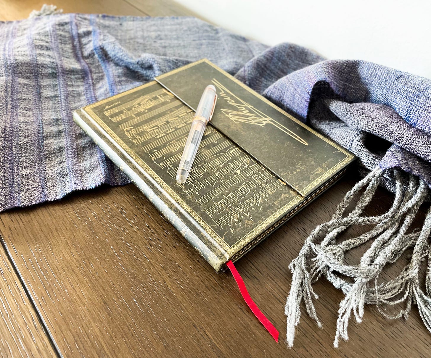 A notebook with music notes on it, a pen sitting on top of it, and a blue shawl around it. All on a brown surface.