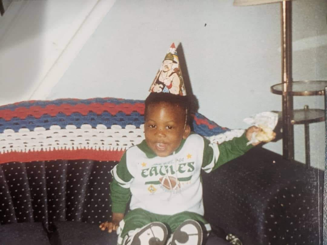 A photo of Nigel when he was 3, wearing Eagles sweatsuit, smiling and sitting on a couch.