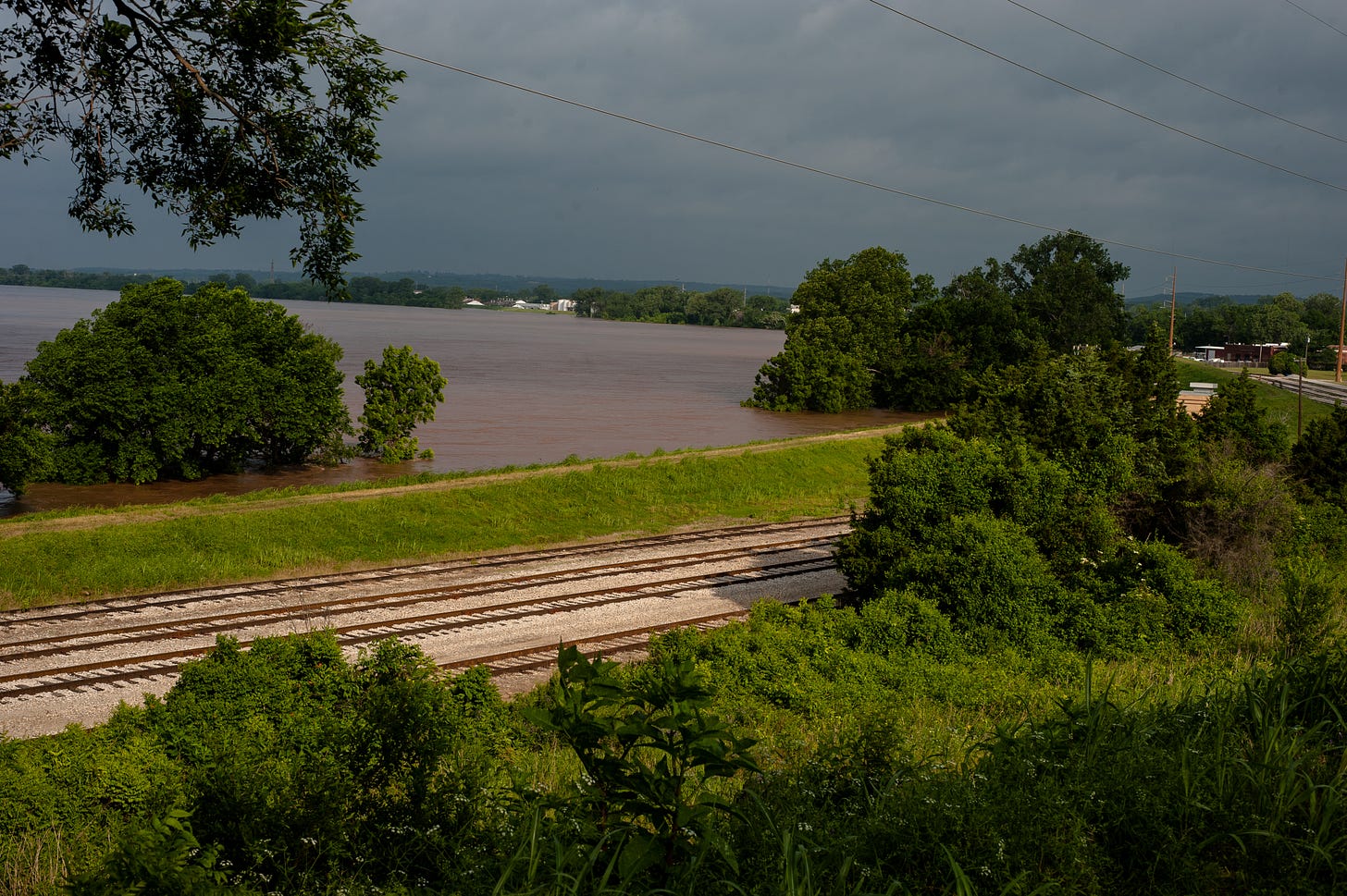 The mud-red Arkansas River submerges large trees to mid-canopy as it rises to within a few feet of the top of a grass-covered levee. The image perspective is from inside the leveed area along a curve, with train tracks in the foreground. A peculiar light hits the levee as clouds hang in the overcast, ominous-looking sky.