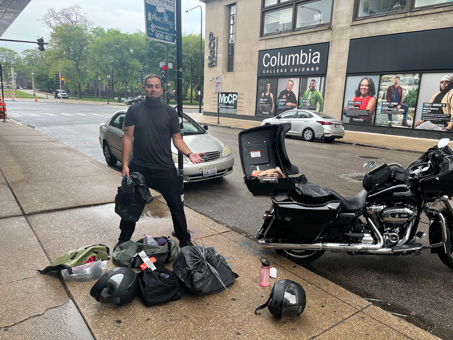 Derrick stands on a sidewalk with all their gear from the motorcycle in front of him. The pavement is wet from rain. The bike is parked on the road next to Derrick. Derrick smiles at the camera.