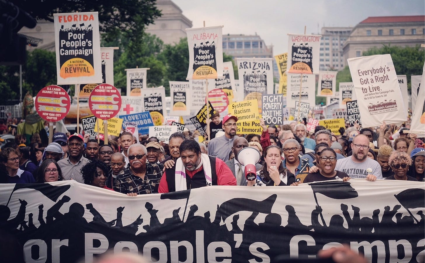 A protest with a big sign at the front that says "Poor People's Movement" with a reverand in his reverand clothes in the middle behind the sign. With a hundred or more people by his side and behind him holding up signs.