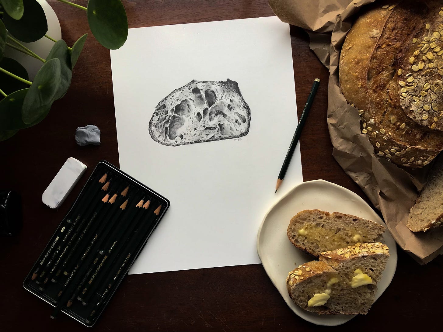 A shot of the finished drawing with a loaf of bread to the right of it, pencils below and a plant to the top left corner. 