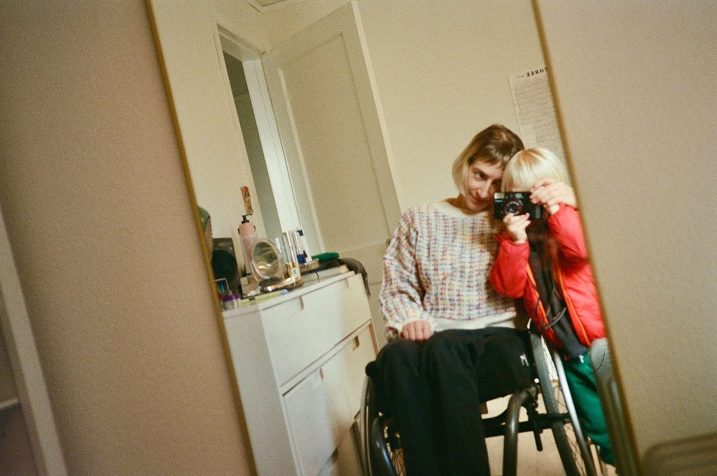 Rebekah sits in her wheelchair next to her son, their heads touching. She is looking toward him as he holds a 35mm camera up to his face, taking a picture of the two of them in a long mirror. Beside them and reflected in the mirror, a cluttered dresser.