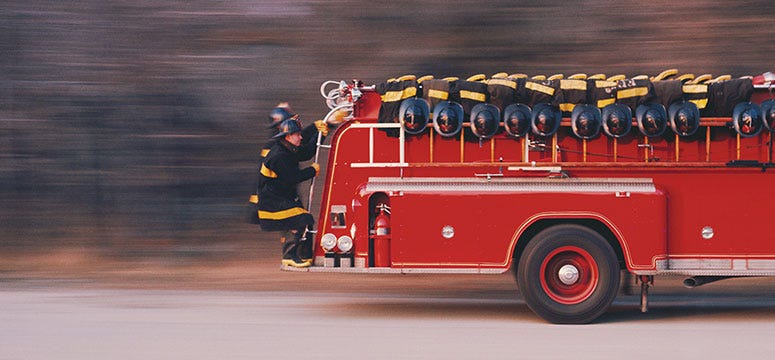 Photo of a model of an old-fashion fire truck with firefighters standing in crouched positions standing on a little ledge that traverses the back of the vehicle, holding onto handles to keep from falling off.