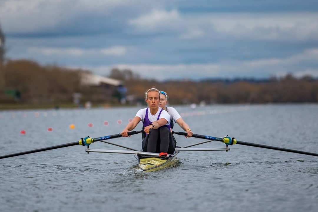 Photograph of rowing boat on a lake, with grey clouds and boat house in the background. Rowing boat has two women in it, each hold an oar. Blonde woman in stroke is wearing white tshirt, black leggings and purple unisuit. Blonde woman in bow also wearing white t shirt - she is mostly hidden. Photograph of rowing boat on a lake, with grey clouds and boat house in the background. Rowing boat has two women in it, each hold an oar. Blonde woman in stroke is wearing white tshirt, black leggings and purple unisuit. Blonde woman in bow also wearing white t shirt - she is mostly hidden.