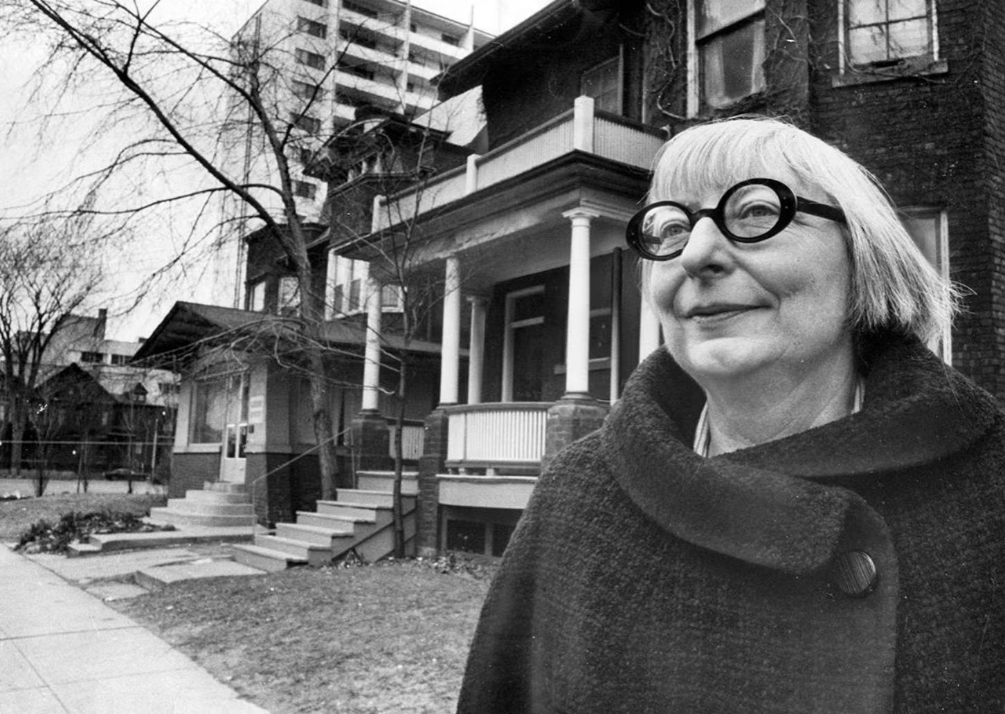 Black and white photograph of Jane Jacobs standing in front of her home. Jane has a winter coat on, a short bob with bangs and is wearing her iconic black framed glasses.