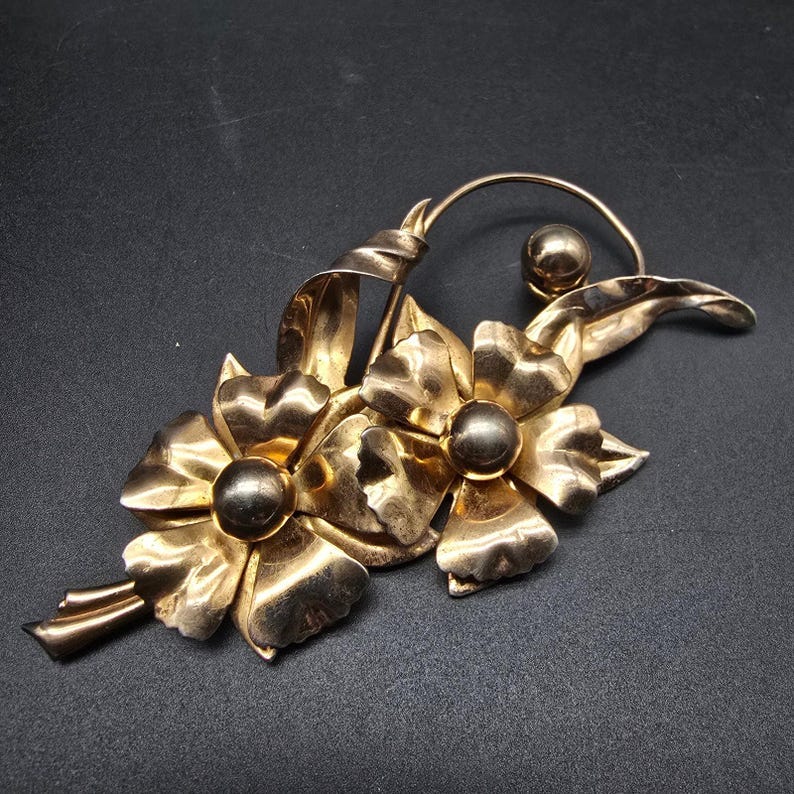 May include: A gold-toned flower brooch with three flowers and leaves. The flowers have a textured surface and each has a round, dark-toned center.