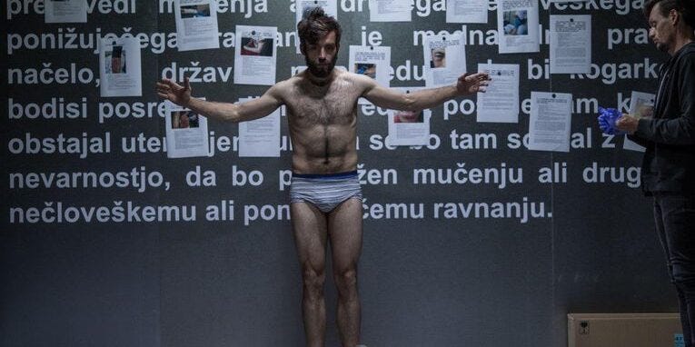 Scene from The Game. A man stands in front of a wall in his underwear. On the wall are testimonies of migrants about their experience of border violence.