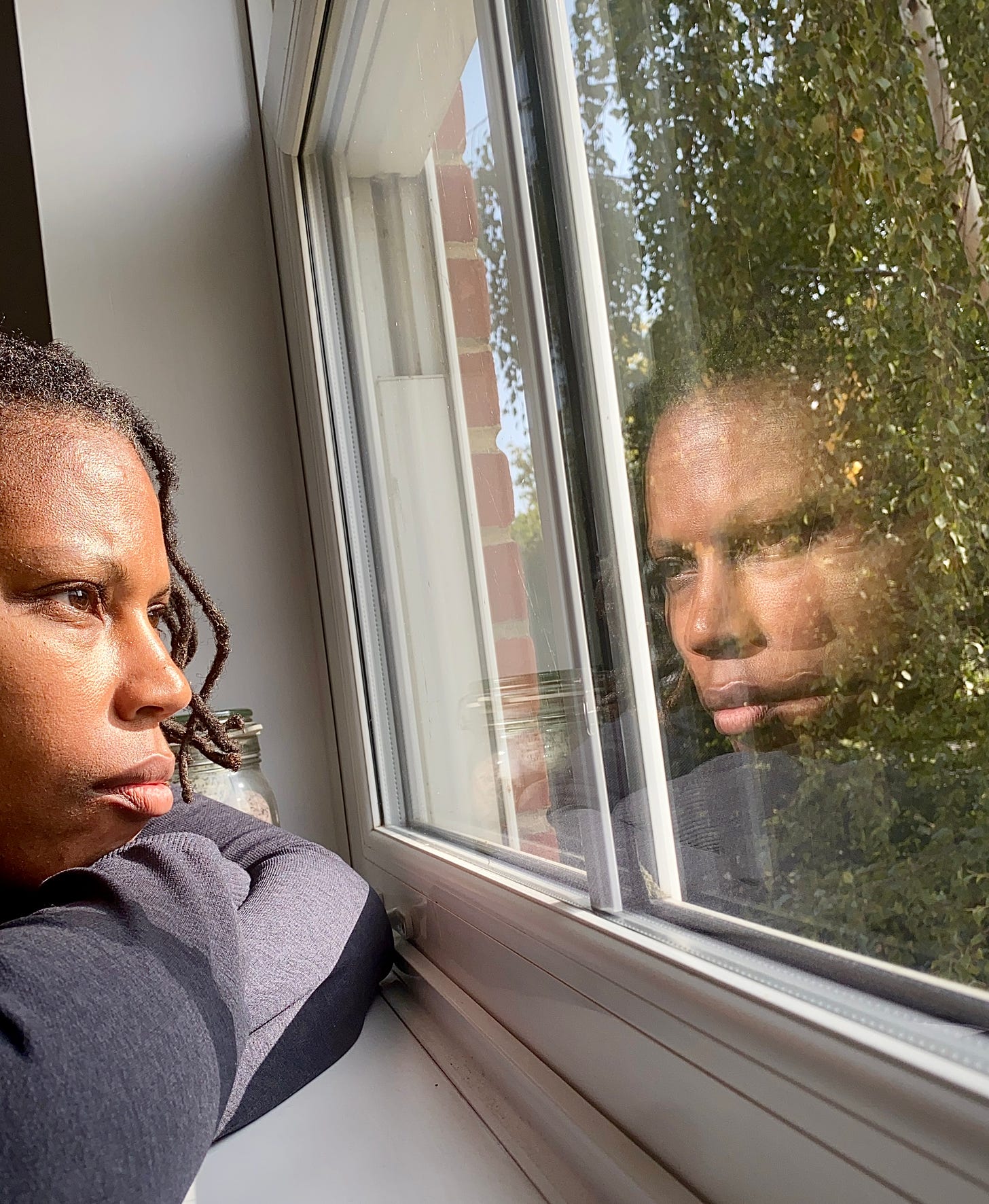 A Black femme stares out the window with their reflection looking back at them