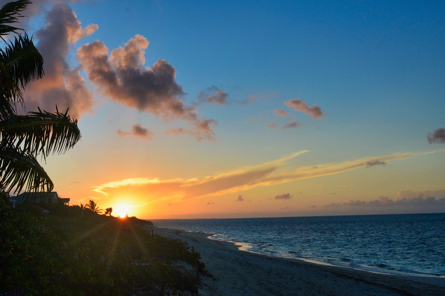 The sun sets on the beach with clouds in the sky on the left with pal fronds on the edge; the sun’s setting rays reach into the blue sky