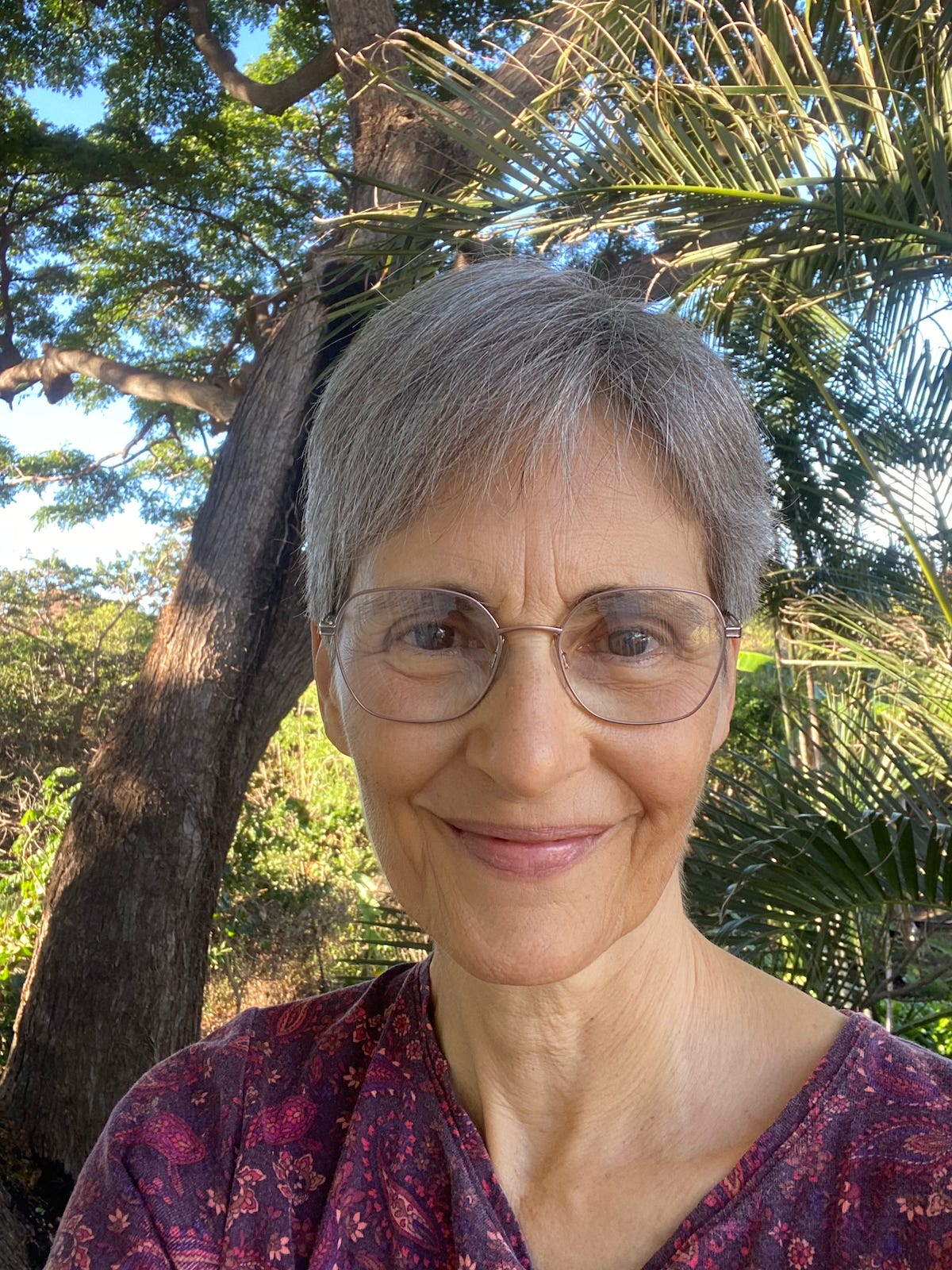 Head and shoulders of a smiling white woman outdoors in front of a large tree and palm fronds.