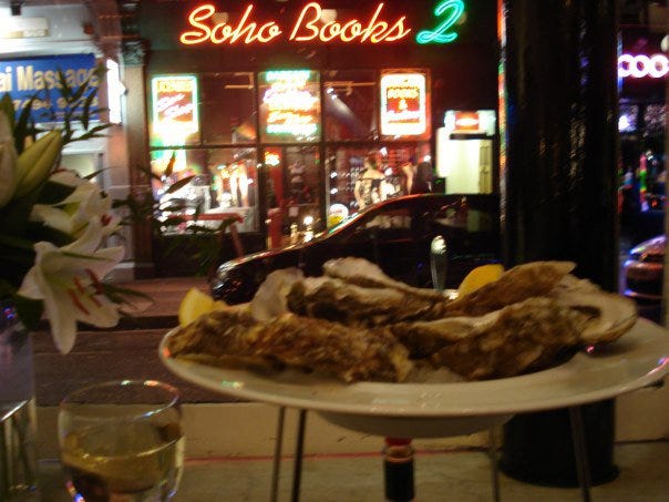 A plate of oysters in a restaurant in Soho. Through the window beyond the oysters, there is an adult bookstore called "Soho Books 2." Beside it there is a Thai massage parlour.
