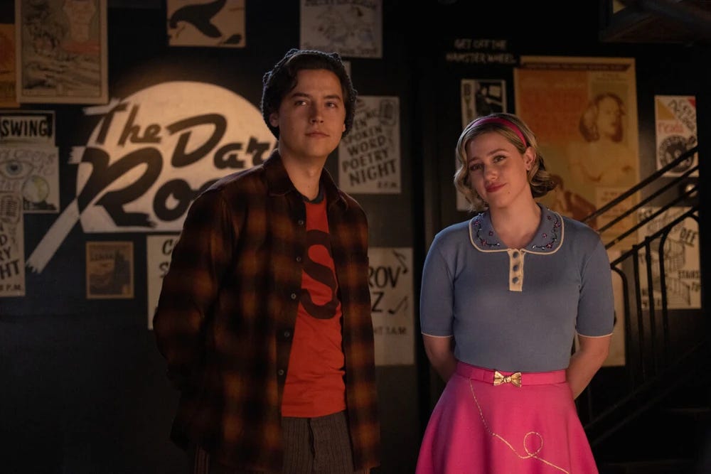 Jughead jones dressed in modern clothes stands next to Betty, dressed in 50s clothes.