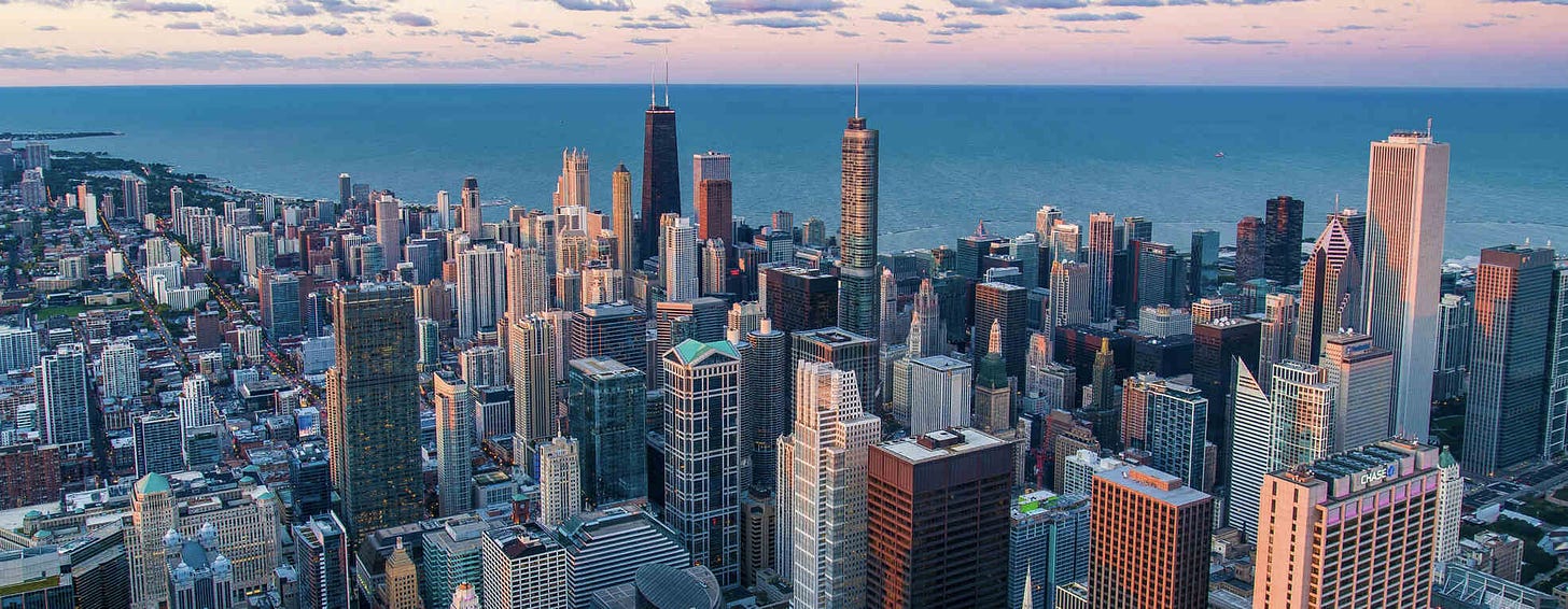 An aerial view of the Chicago skyline at dusk, featuring a mix of high-rise buildings and skyscrapers with Lake Michigan in the background. The sky is painted with soft hues of pink and blue, adding a serene backdrop to the bustling cityscape.