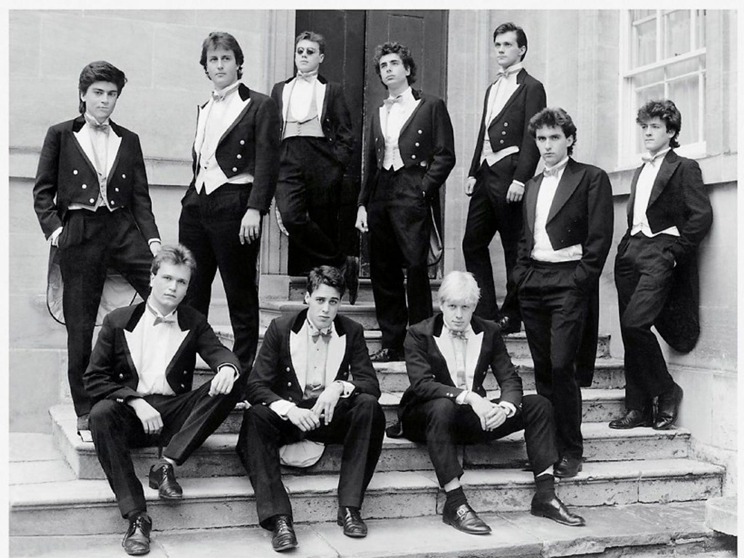 Mid 1980s black and white photo of Oxford University's Bullingdon Club, 10 white men in white tie suits pose poshly on a staircase in front of some double doors.