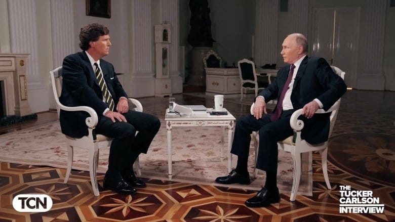 Two men in suits, sitting in white armchairs, face each other in an interview.