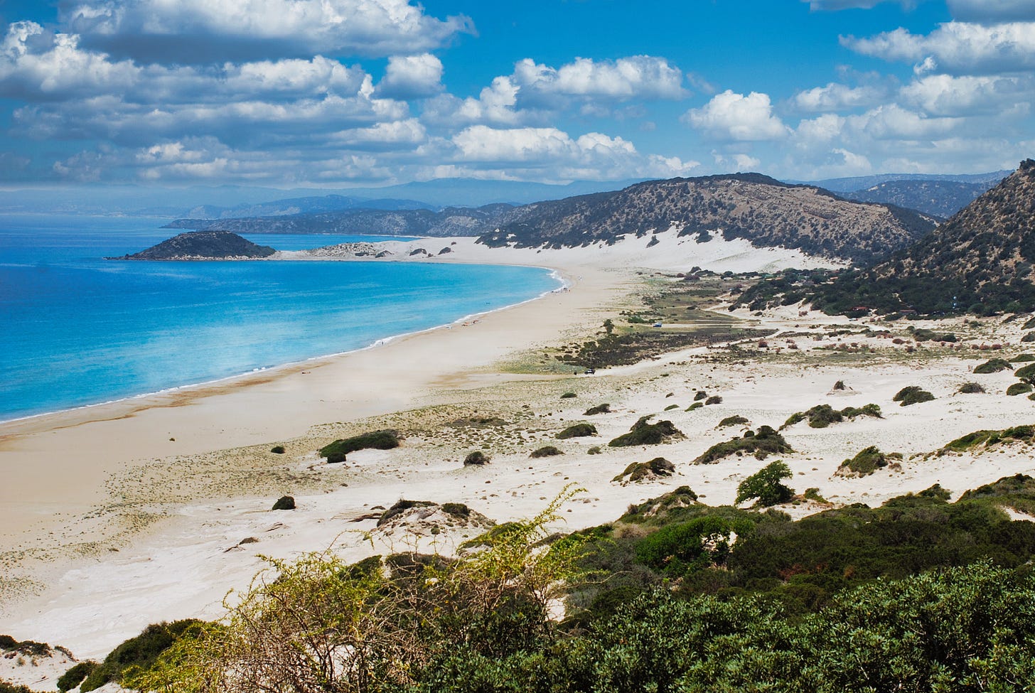 Panoramic view of the long, sandy beach on the Karpas Peninsula in Northern Cyprus, bordered by clear blue water and rolling hills.