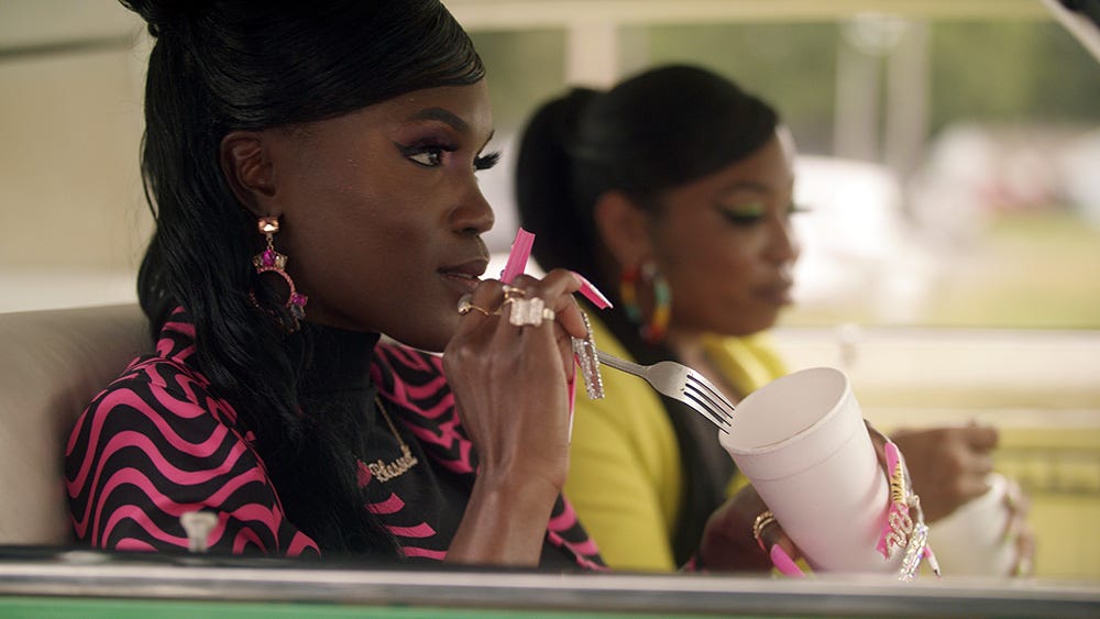 Two fly Black women sit in a car holding styrofoam cups and forks with ramen in them.