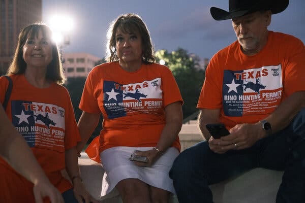 Three people wearing red T-shirts in support of Ken Paxton are shown sitting outside the Texas Capitol Building early Tuesday. One is wearing a large cowboy hat. 