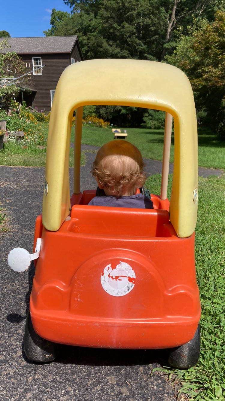 a toddler with blonde curly hair sits in a plastic car. The car is yellow and orange and is on green grass.