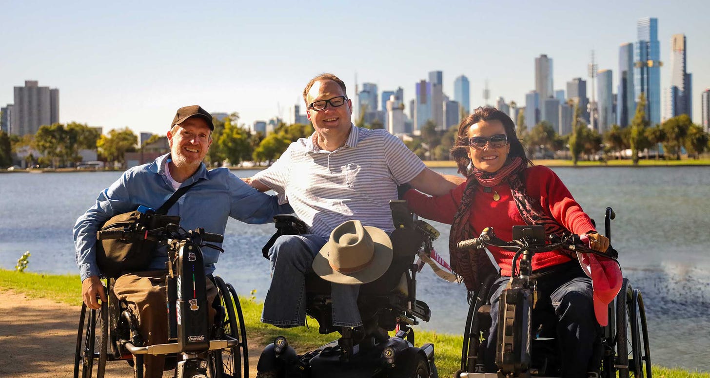 John seated in his wheelchair along with two other wheelchair users in front of the Melbourne city skyline, seen from a park with a large lake.