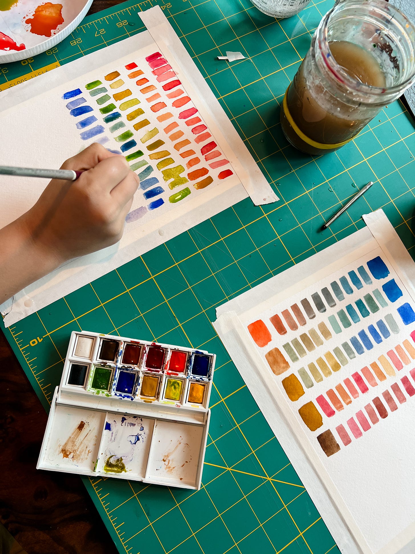 Child's hand holding watercolor brush while making watercolor swatches in a rainbow of colors.