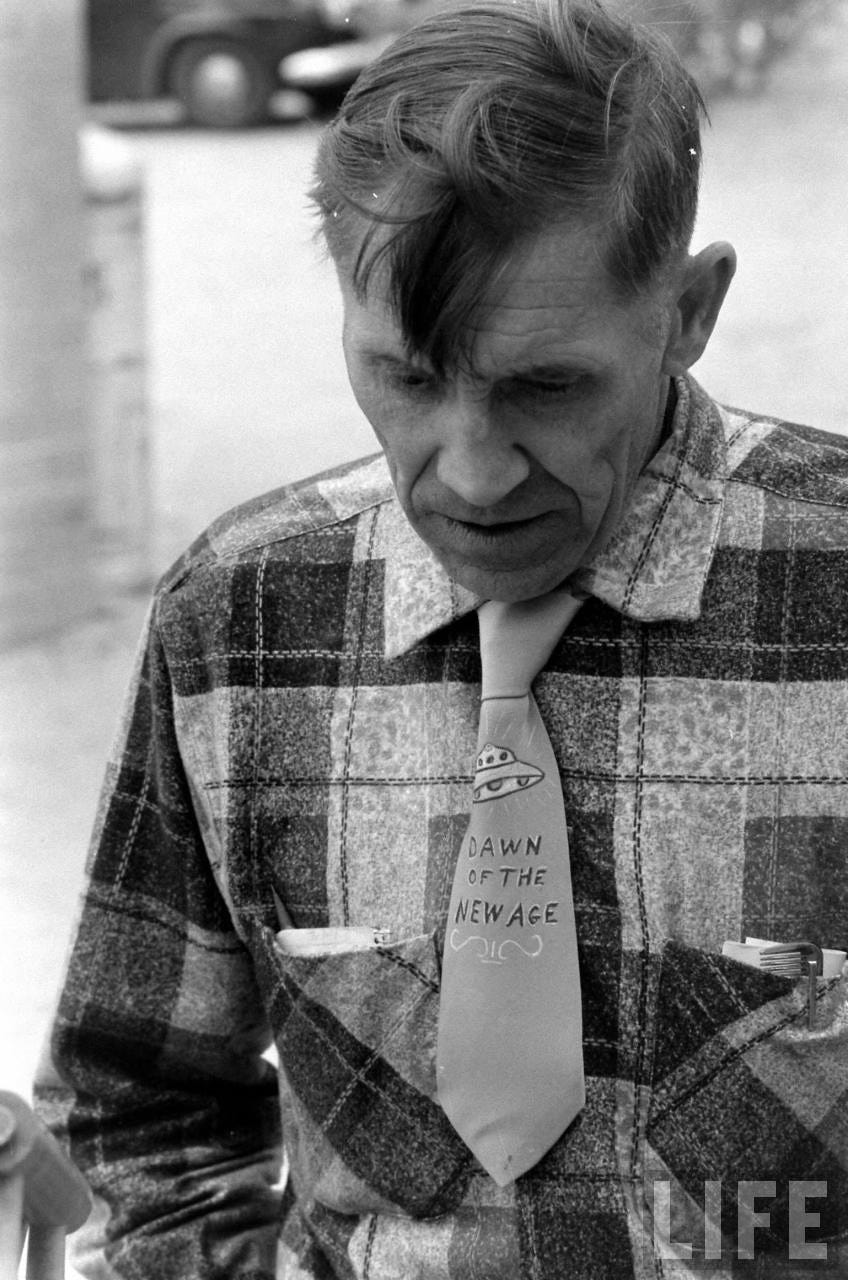 A black and white image of a man in late 1950s clothing, wearing a tie that says "Dawn of a New Age" with a UFO embroidered above the letters.