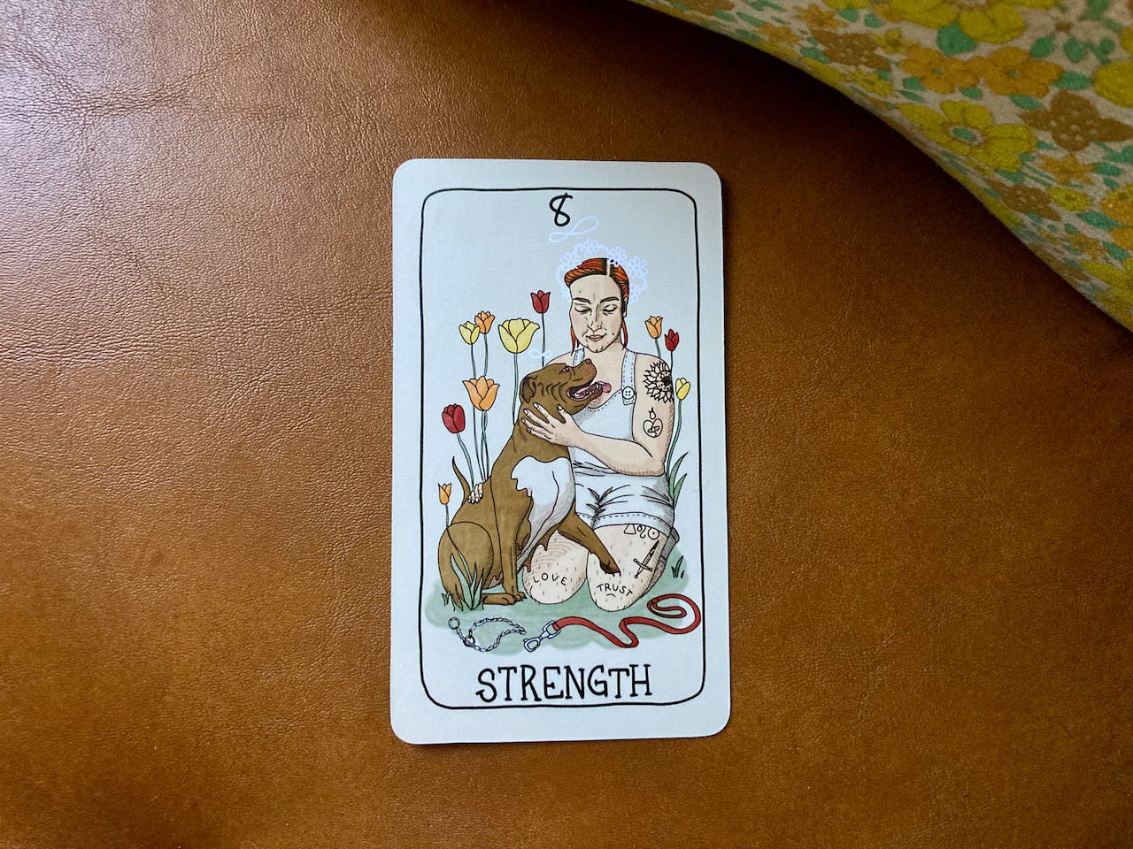 A Strength tarot card sits on a brown leather chair. The card shows a light-skinned person with dyed red hair kneeling and petting a pit bull, who licks them. The dog's choke collar and leash are off and lay on the ground. The person has "love" and "trust" tattooed on their knees.