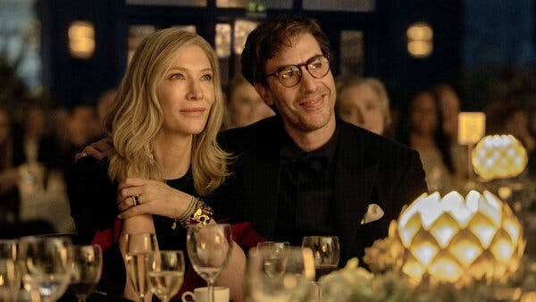 A woman and man sit at a fancy dinner table and smile at something they’re watching.