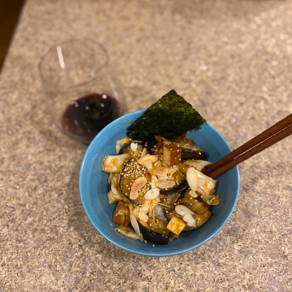 A blue bowl of white rice and the stir-fry described above, topped with toasted almonds and sesame seeds, and a sheet of gim at the edge of the bowl. A pair of wood chopsticks sticks out on the right hand side, and there is a glass of red wine above and to the left.
