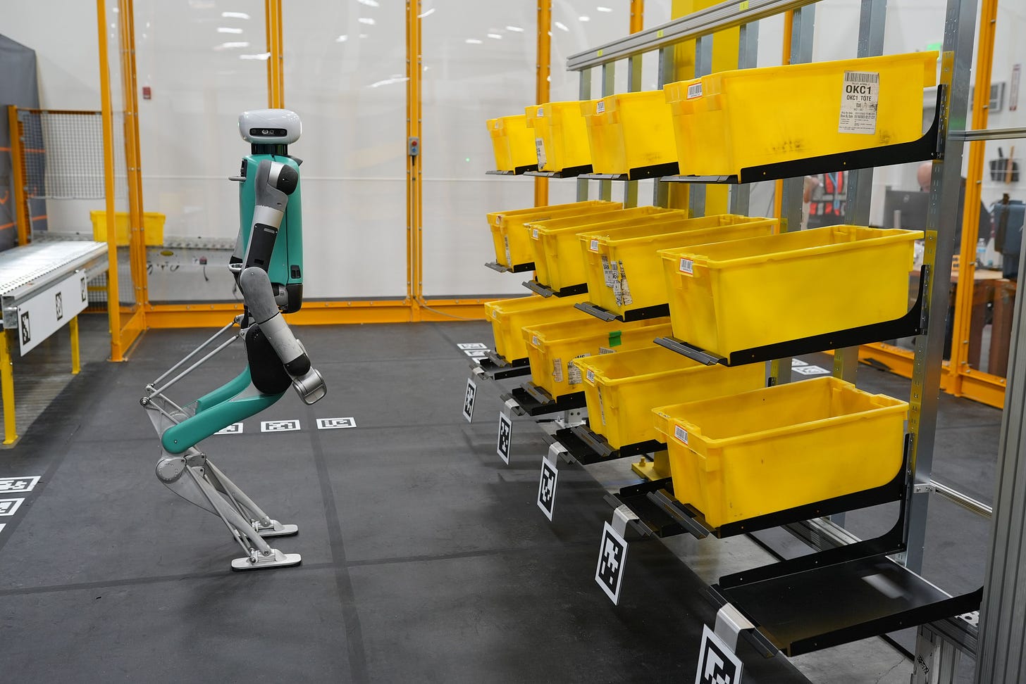 A humanoid robot stands in front of a series of yellow bins for hauling materials.
