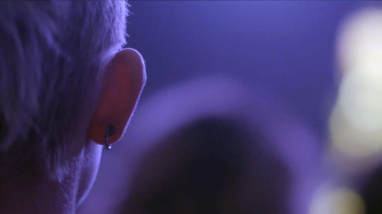 A shot from behind a person standing in purple hues, showing their head and bent barbell earring on the left of the image.