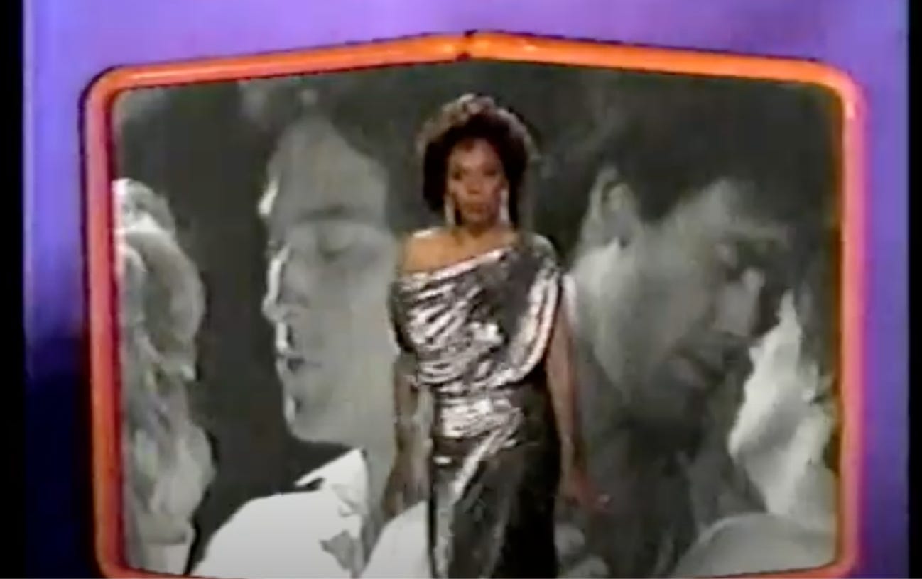 Jenifer Lewis is wearing a silver dress. She’s African American and has short black hair. In the background is two couples kissing. They are pictured in black and white.