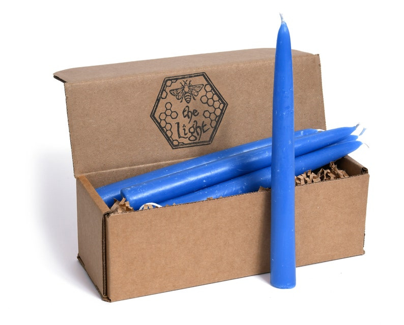 May include: A box of blue taper candles. The box is brown cardboard with a bee and honeycomb logo that reads 'be the light'. The candles are arranged in the box with one candle standing upright outside the box.