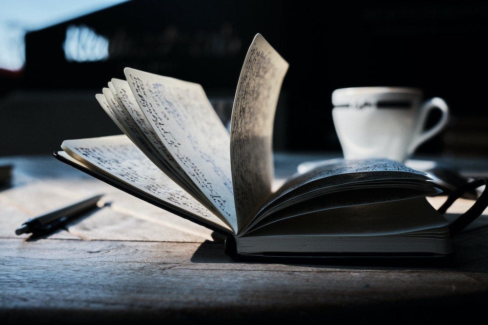 An open notebook beside a pen and a coffee cup on a wooden table.