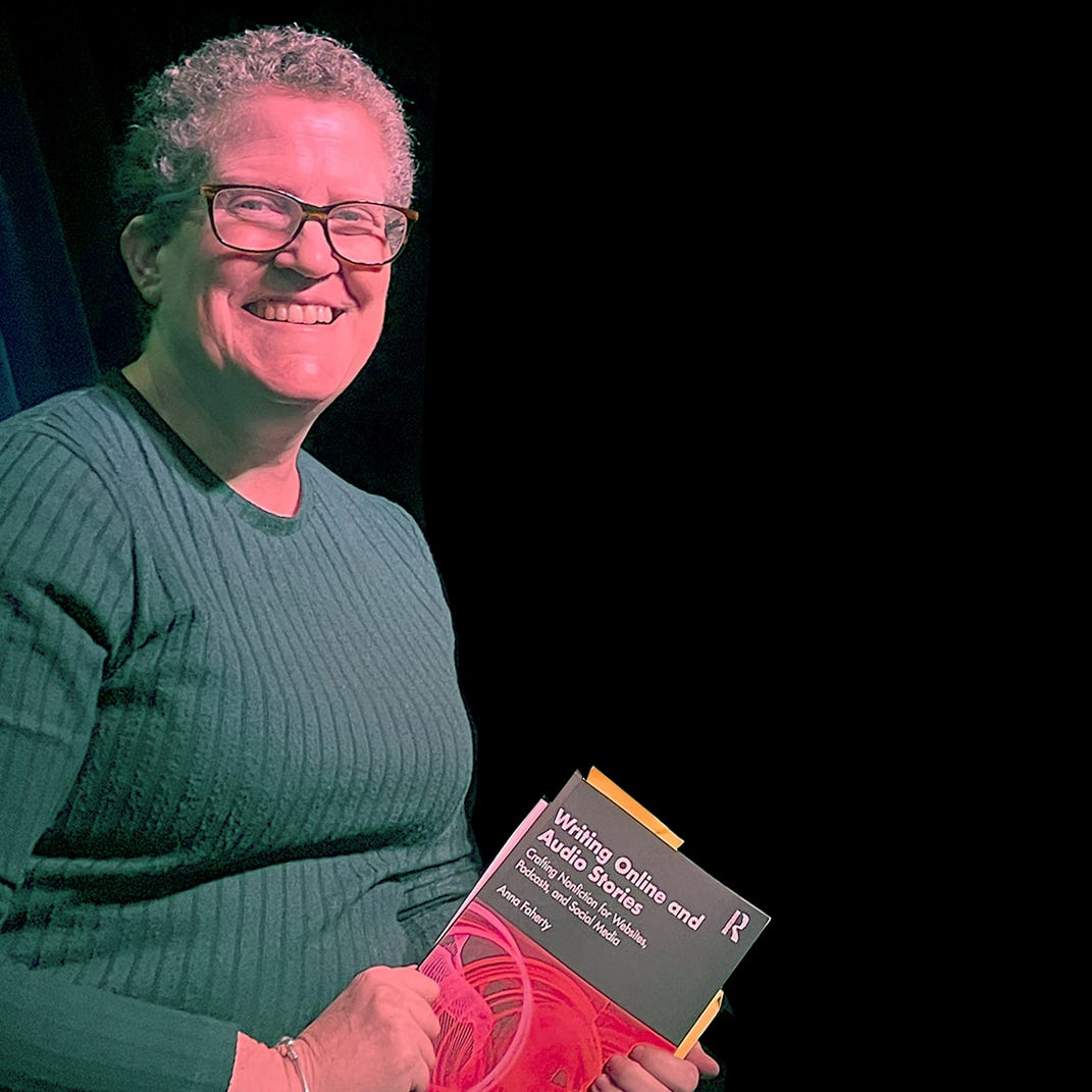 Anna Faherty, a white woman in her 50s with fair skin, short curly hair and glasses. She sits in front of a black background wearing a green jumper and holds a book in her hands.